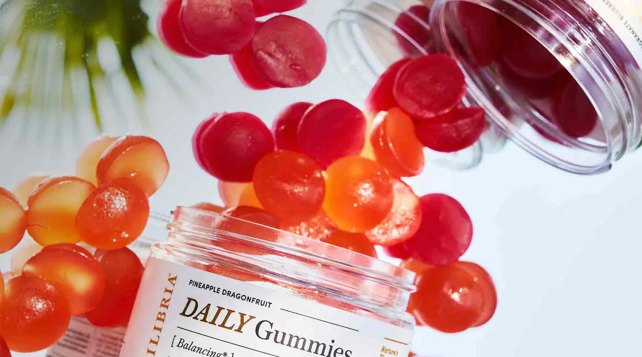 A closeup photo of Equilibria's vibrant red CBD gummies spilling from one jar to another on a white background with some foliage blurred out in the background.