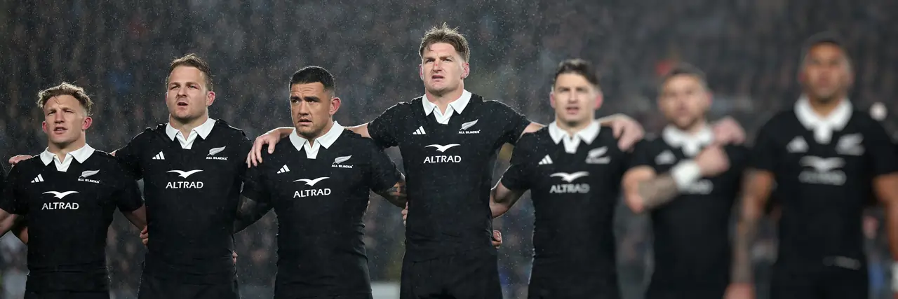 The New Zealand AllBlacks rubgy team stand triumphantly in the rain with the crowd blurred out in the background.