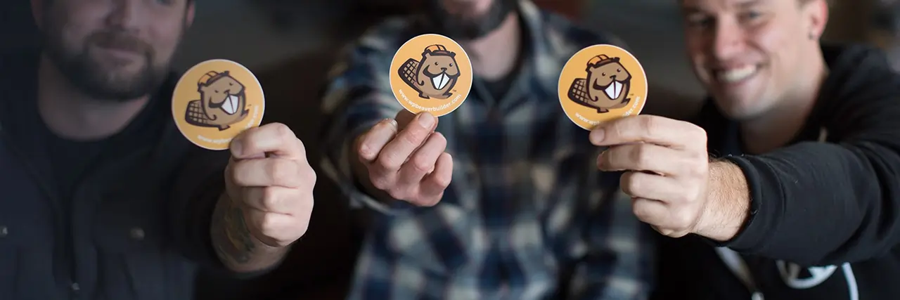 Three men from the Beaver Builder team hold up stickers of their beaver logo to the camera, with a shallow depth of field so their bodies are slightly out of focus.