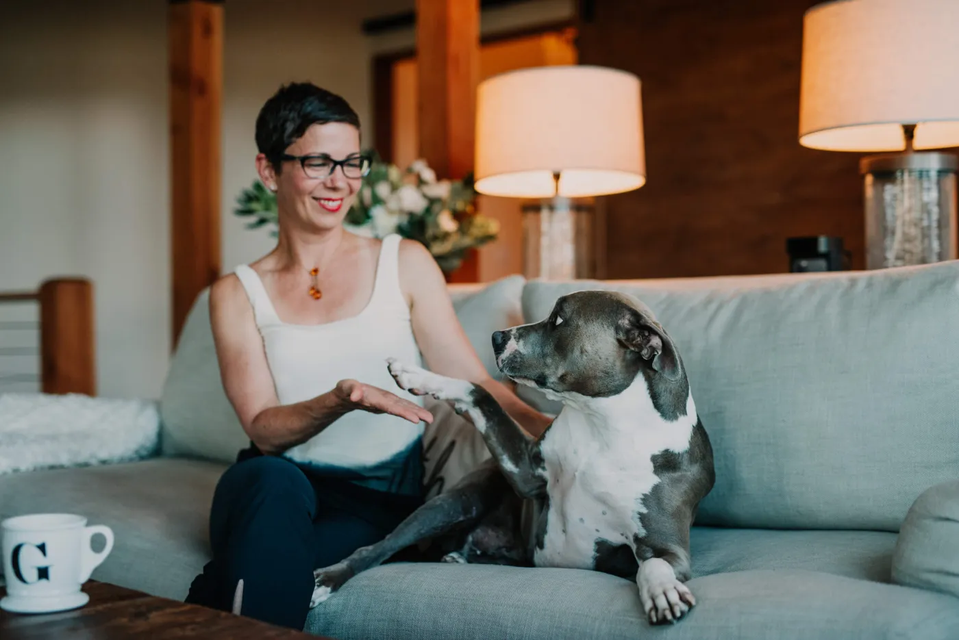 Greta Rose Zagarino smiling on a couch and offering her hand to her grey and white dog, a mug with a G on the coffee table.