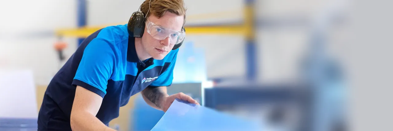 Online Plastics Group: a technician in safety glasses and ear defenders handling a sheet of blue plastic in the workshop.