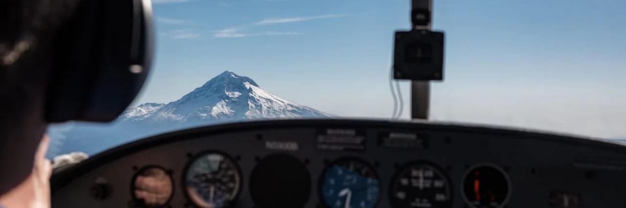 From behind the pilot of a small plane you see the pilot and his headset in the foreground with the rising alps in the background.