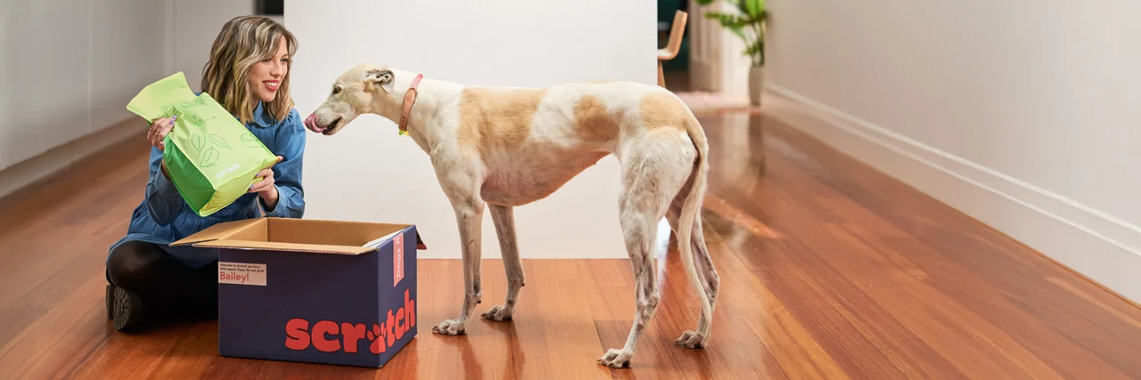 A blonde woman in her 30s sitting on the timber floor in the kitchen of her modern apartment lifts a lime green bag of Scratch Pet Food out of a navy blue shipping box whilst her white greyhound dog licks its lips, hungry for dinner.