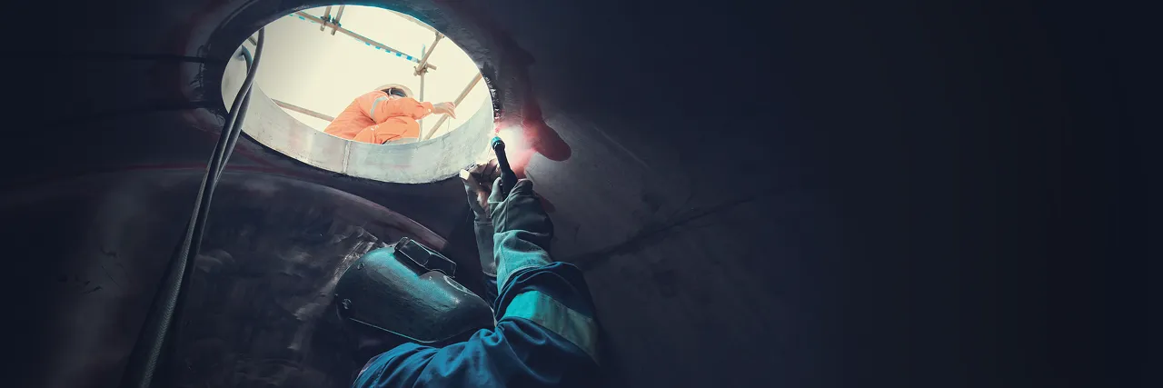 Start Training: a welder at work inside a steel tank, lit by the torch, while a supervisor in orange overalls watches through the hatch above.